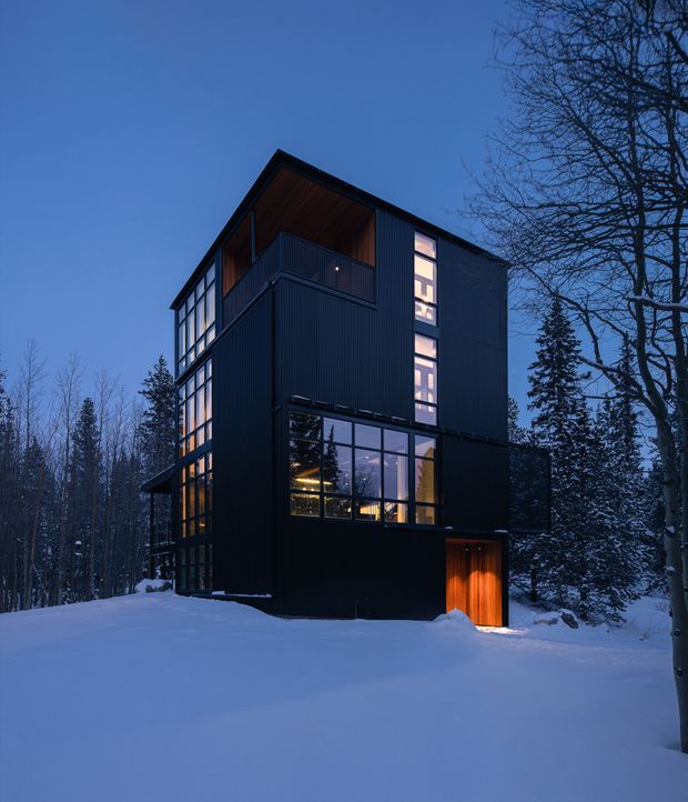 A tall, narrow contemporary house set within a snowy forest at dusk. The building has dark vertically ribbed cladding, extensive floor-to-ceiling glazing, a warm timber-lined recessed upper terrace, and a glowing timber entry door. Interior lights create a warm contrast against the cool blue snow and surrounding bare and evergreen trees.
