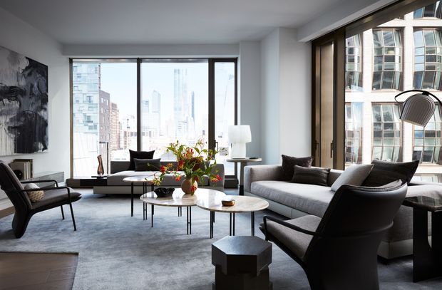 A bright contemporary high-rise living room with expansive corner glazing and city views. The room features a large pale sectional sofa, several dark leather lounge chairs, nested sculptural coffee tables, a soft grey rug, minimalist lighting, and abstract wall art.