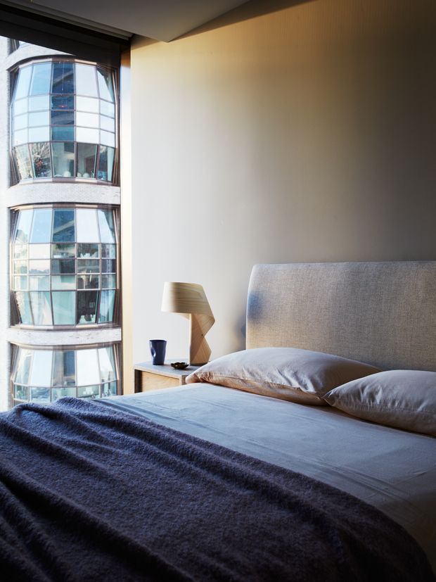 A calm contemporary bedroom with a large upholstered bed, pale bedding, and a dark textured throw. A small wooden bedside table holds a sculptural lamp and dark cup. Floor-to-ceiling glazing provides an urban view of a curved glass-fronted building, while warm neutral wall finishes and soft daylight create a refined atmosphere.