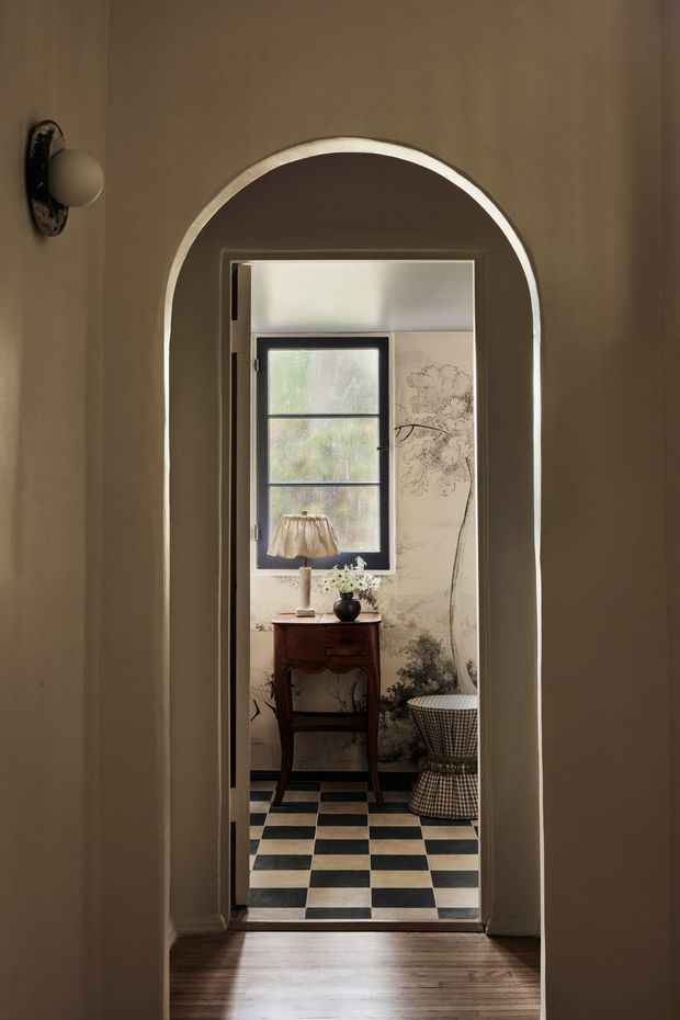 A moody interior view through a rounded archway into a compact, traditionally styled bathroom or powder room. The room features a black-and-cream checkerboard tile floor, botanical wall covering, a dark timber console vanity with a small lamp and decorative objects, a black-framed window, and a patterned upholstered stool. The foreground has warm timber flooring, taupe walls, and a vintage wall sconce.