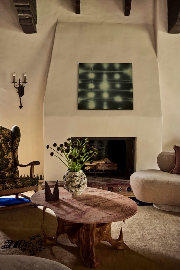 Warm, richly styled living room with a tall sculpted plaster fireplace wall, exposed dark timber ceiling beams, and a recessed open fireplace. The room features a rounded beige sofa, ornate patterned lounge chair, sculptural wood coffee table with a mottled stone-like top, textured rug, decorative vase with dried seed pods, wall art, and a wrought-iron candle sconce. The overall aesthetic is rustic, eclectic, and refined with earthy tones and dramatic natural light.