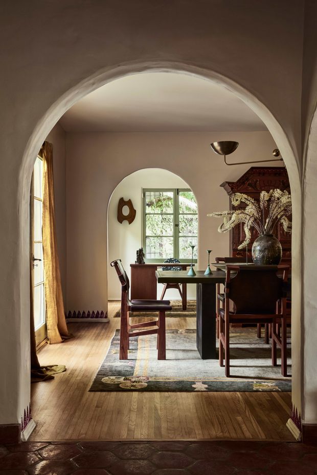 A warm, richly layered dining room viewed through a broad plastered archway. The room features a dark timber dining table and chairs, a large ornate dark wood cabinet, patterned rug, timber flooring, arched niches and openings, a central pendant light, decorative ceramics, and dried floral arrangements. Natural light enters through tall windows with ochre curtains, creating an intimate, historic-feeling interior with strong Mediterranean and traditional character.