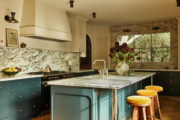 A richly detailed traditional kitchen with a large marble-topped island, deep teal-blue cabinetry, brass hardware, a classic range cooker, and a substantial plaster rangehood. A dramatic marble backsplash runs along the cooking wall, while exposed brick surrounds a large black-framed window. Timber flooring, warm neutral cabinetry, vintage-style lighting, and curved timber stools create a refined, warm atmosphere.