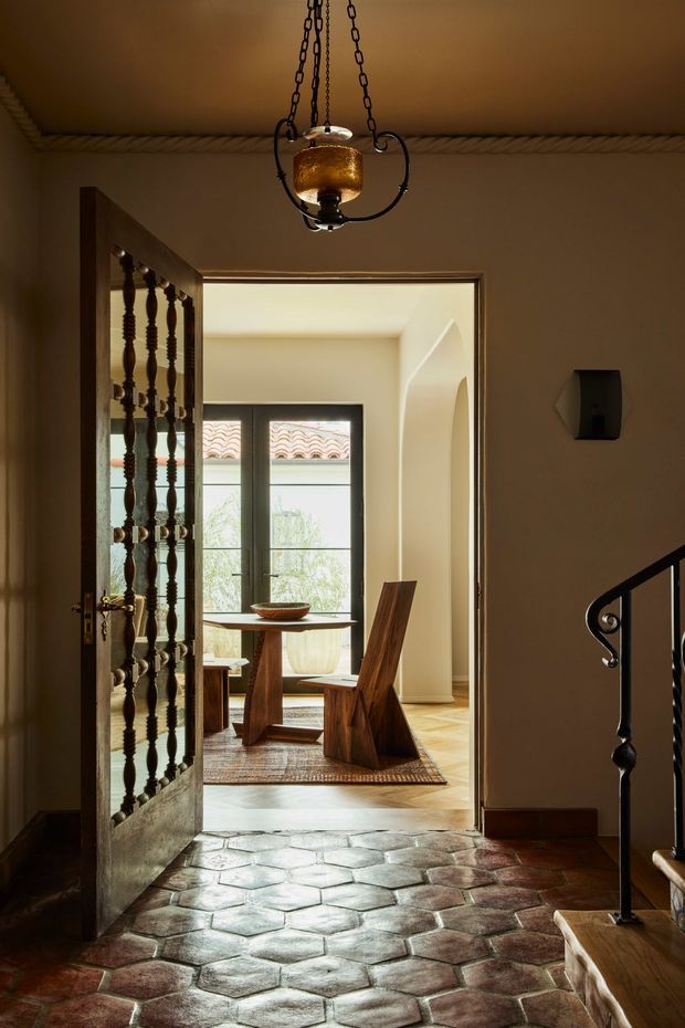 A warm, moody entryway viewed toward a bright dining room. The foreground has irregular terracotta-toned hexagonal tile flooring, dark wood trim, a decorative timber-and-metal open door, and a black wrought-iron stair balustrade. A hanging amber glass lantern illuminates the ceiling. Through the doorway, a timber dining table and sculptural wooden chairs sit on a woven rug, with dark-framed windows and an arched wall detail.