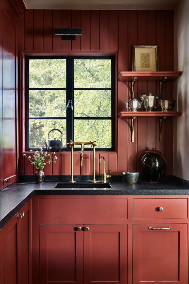 A richly coloured kitchen scullery or wet-bar area with deep red painted vertical wall panelling and matching shaker-style cabinetry. A black stone benchtop surrounds a compact undermount sink with brass tapware, positioned beneath a black-framed divided window overlooking leafy trees. Open red shelves display trophies, framed artwork and decorative vessels, while dark finishes and warm lighting create a moody, refined atmosphere.