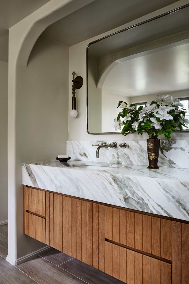A refined bathroom vanity area with a long floating timber-grain cabinet, a thick sculptural white marble benchtop and matching marble backsplash, wall-mounted metal tapware, and a large rounded-corner mirror. The room has softly curved architectural detailing, warm neutral painted walls, dark timber-look flooring, and a decorative wall sconce. A floral arrangement sits on the vanity.