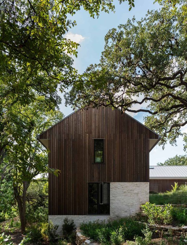Exterior view of a contemporary gabled house set among mature trees and landscaped planting. The facade combines dark vertical timber cladding with a pale textured masonry or stone base, narrow black-framed windows, and a simple pitched roof. A secondary wing with a light metal roof is visible behind the main volume.