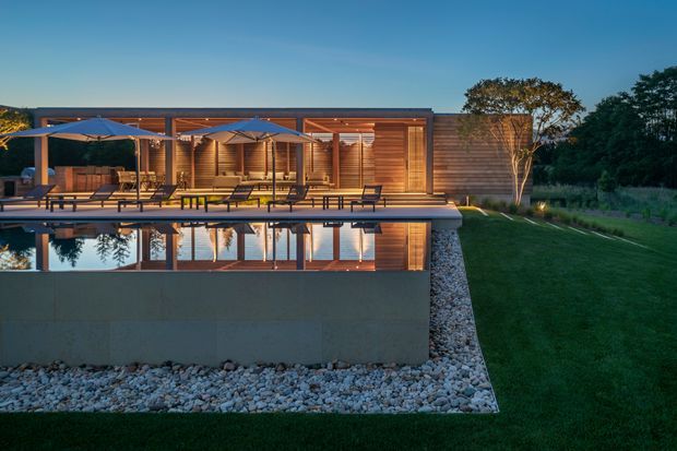 Twilight exterior view of a low-slung contemporary timber-clad residence beside a reflecting swimming pool. The building has extensive glazed openings, a covered timber deck with outdoor lounge furniture and umbrellas, warm interior lighting, and a landscaped lawn with a planted tree. A concrete pool structure and decorative rock bed frame the foreground.
