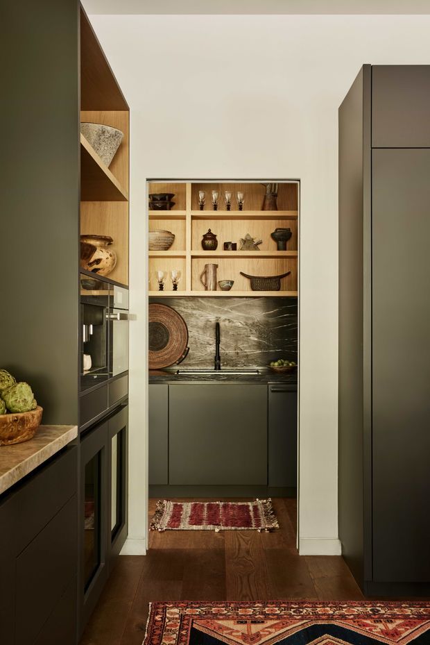 A warm, refined kitchen view looking through a narrow opening into a compact secondary kitchen or butler’s pantry. The main kitchen has dark matte cabinetry, integrated appliances, timber flooring and a stone benchtop. The back space features dark lower cabinets, an open timber display shelf filled with glassware and ceramics, a dramatic dark marble splashback, and a brass-toned tap. Patterned woven rugs and curated objects add texture and character.