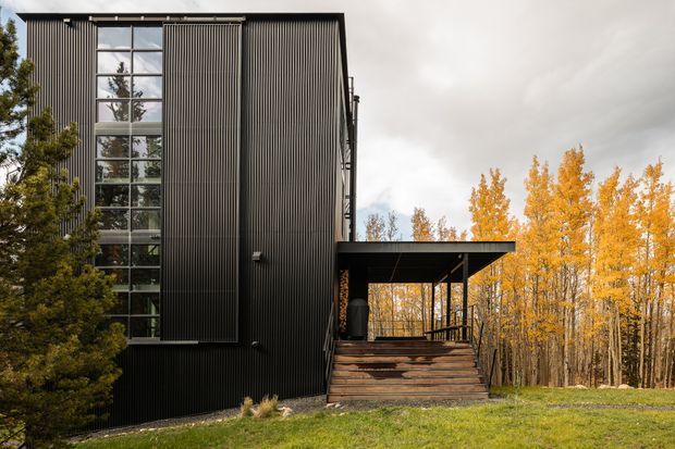 Exterior architectural photograph of a tall, narrow modern building clad in dark vertically ribbed metal, with a large multi-level glazed window wall and a projecting covered entry deck reached by broad timber steps. The building is set among mature trees with vivid autumn foliage and a grassy clearing under an overcast sky.