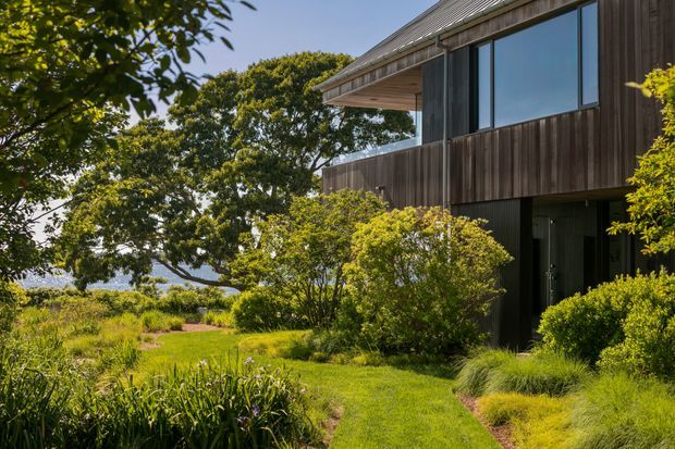 Exterior view of a contemporary two-storey residence set within a lush landscaped garden. The building features dark vertical timber cladding, broad glazed openings, a projecting upper floor with a timber-lined soffit, and a low-pitched roof. A grassy path leads through dense ornamental planting toward the recessed entry.