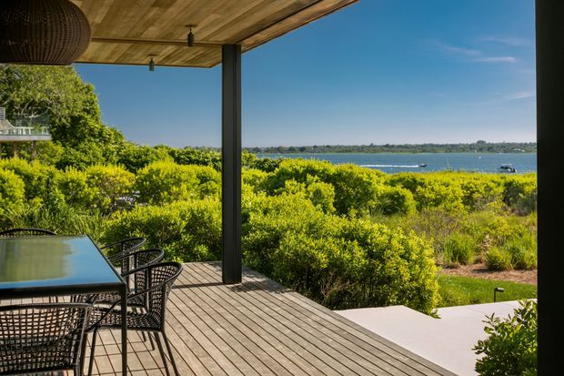 A covered timber deck overlooking dense coastal vegetation and a broad body of water. The outdoor dining area includes a glass-topped table with woven black chairs, a timber-lined ceiling, dark structural posts, and a large woven pendant light. Steps lead down toward a pale paved path and landscaped garden, with a bright blue sky and distant shoreline beyond.