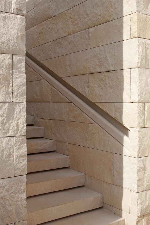 Close-up architectural photograph of a compact stairway clad in pale beige natural stone. Thick rectangular stone blocks form the walls and stair treads, with a matching integrated stone handrail running diagonally along the side. Strong sunlight creates warm highlights and shadows, emphasizing the textured masonry and clean geometric lines.