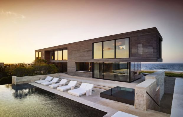 Modern two-storey coastal residence with a dark horizontal timber-clad facade, extensive floor-to-ceiling glazing, and a large outdoor terrace overlooking the ocean. The terrace includes a swimming pool, sun loungers, and a sunken dark-water spa or feature pool, with pale stone paving and retaining walls.
