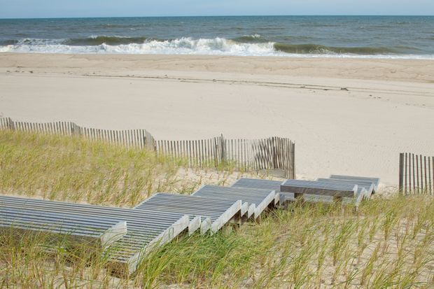 A coastal beach landscape viewed from above, with pale sand, rolling ocean waves, dune grasses, timber sand fencing, and a stepped grey-toned boardwalk leading toward the beach.