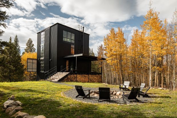 A striking contemporary black-clad multi-storey house set in a wooded landscape, with tall glazing, an elevated entrance stair, projecting flat-roofed canopy, and a gravel outdoor seating area with a fire pit in the foreground. Golden deciduous trees and evergreen vegetation surround the home.