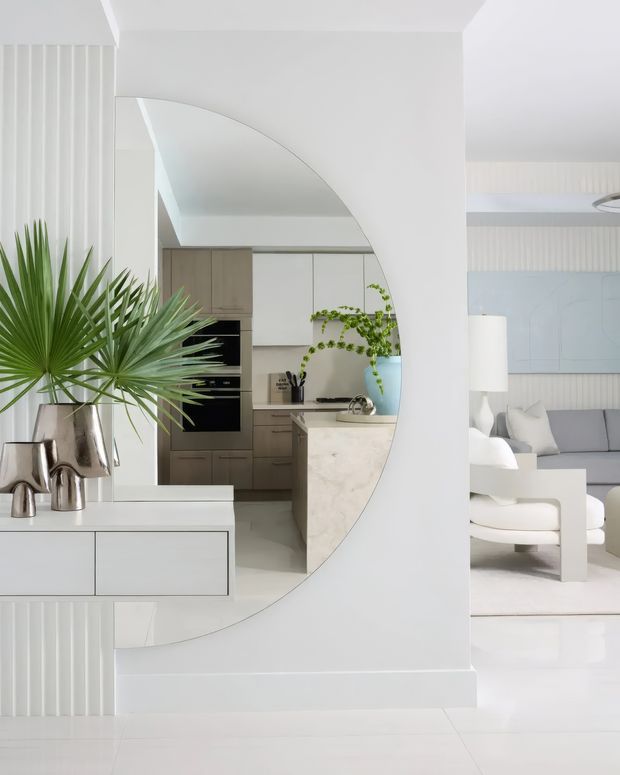 A bright, refined open-plan interior with a large arched full-height mirror reflecting a compact kitchen. The foreground features a white built-in console with sculptural metallic vessels and tropical foliage, while the adjoining living area includes pale upholstered seating, a white coffee table, and a soft blue wall artwork. Vertical white wall panelling and predominantly white finishes create an airy, minimalist composition.