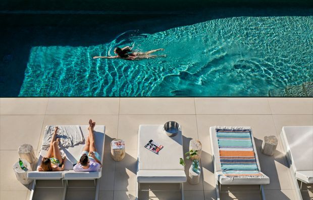 A top-down view of a swimming pool with a swimmer, alongside a tiled pool deck furnished with white sun loungers, striped towels, drinks, and small potted plants. The scene has a resort-like outdoor leisure setting with strong sunlight and turquoise water.