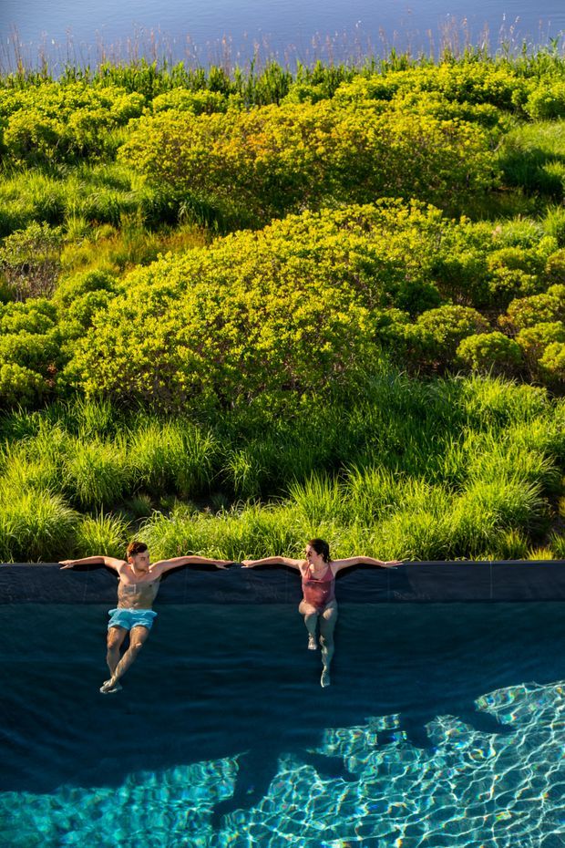 A close view of a swimming pool integrated into a lush natural landscape. Two people are resting at the dark pool edge, with clear blue water in the foreground and dense green grasses, shrubs, and reeds behind them. A calm blue body of water is visible in the distant background.