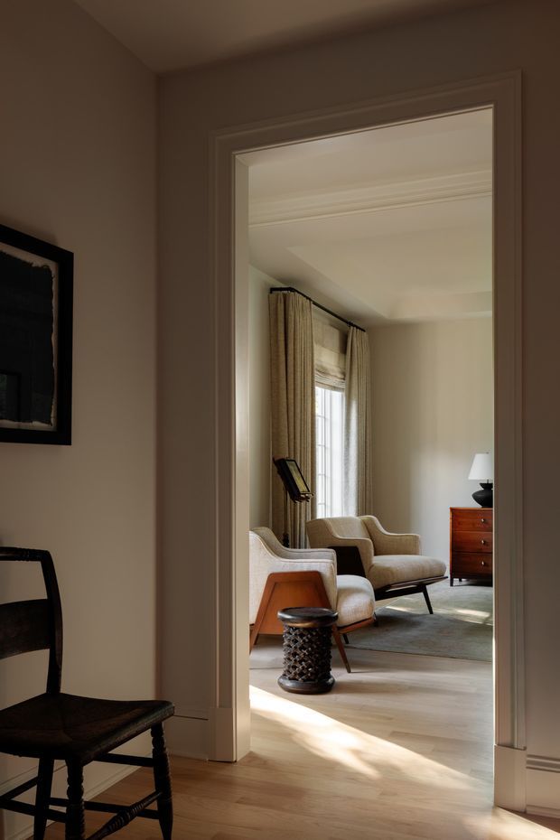A warmly lit view through a wide, traditionally trimmed doorway into a refined living room. The room features upholstered mid-century-style armchairs, a timber chest of drawers, floor-length curtains, a table lamp, a textured area rug, and pale timber flooring. The foreground includes a dark wooden chair and framed wall artwork. Architectural details include substantial door casing, decorative ceiling moulding, and a muted neutral palette.