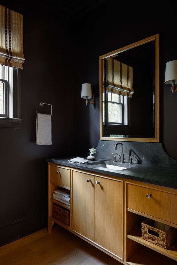 A moody, refined bathroom vanity with dark charcoal walls, warm timber cabinetry, and a dark stone countertop with integrated double basins. A large brass-framed mirror reflects the window and patterned Roman blind. Brass wall sconces, brushed metal tapware, a towel rail, woven storage baskets, and timber flooring complete the traditional yet updated interior.