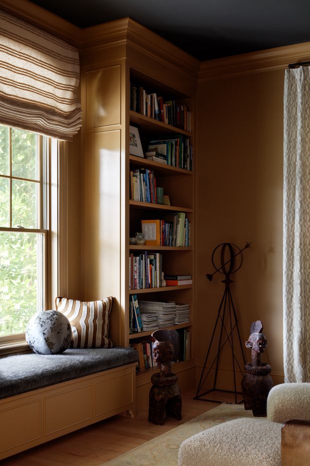 A warm, richly detailed reading nook or study corner with built-in timber cabinetry and open bookshelves surrounding a window seat. The room features honey-brown wood panelling and joinery, a dark painted ceiling with substantial crown moulding, woven Roman blinds and curtains, a sheepskin-textured window seat and armchair, decorative sculptures, books, and a soft neutral rug. Natural daylight enters through the window, creating a cosy, moody and refined atmosphere.