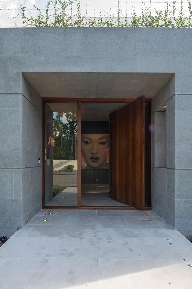 A dramatic contemporary residential entrance framed by large grey concrete or stone wall planes and a deep recessed portal. The entry has a warm timber-framed glazed door assembly with a tall pivot door, concealed floor-level lighting, and a polished grey tiled or concrete approach. A large artwork is visible inside the dark entry hall, while garden foliage is visible through the glazing and above the wall.