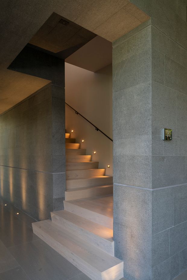 A dramatic contemporary interior stairway with broad pale timber or stone treads ascending through a dark, angular opening. The surrounding walls and floor are clad in large-format grey stone or concrete-like panels, with warm recessed step lights and a slim black handrail creating a refined, moody atmosphere.