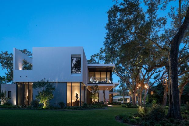A dusk exterior view of a contemporary multi-level residence set within a landscaped garden. The house features crisp white and dark grey geometric volumes, extensive floor-to-ceiling glazing, projecting upper-level balconies, and warm interior and landscape lighting. Mature trees frame the property, with a broad lawn in the foreground.