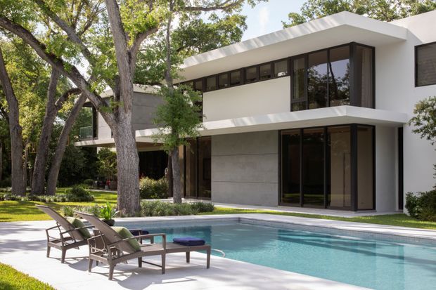 Exterior view of a two-storey modern residence beside a swimming pool, featuring crisp white rendered walls, broad horizontal roof and balcony overhangs, dark-framed floor-to-ceiling glazing, and a large smooth grey feature wall. Mature trees and landscaped planting surround the pool terrace, with outdoor loungers in the foreground.