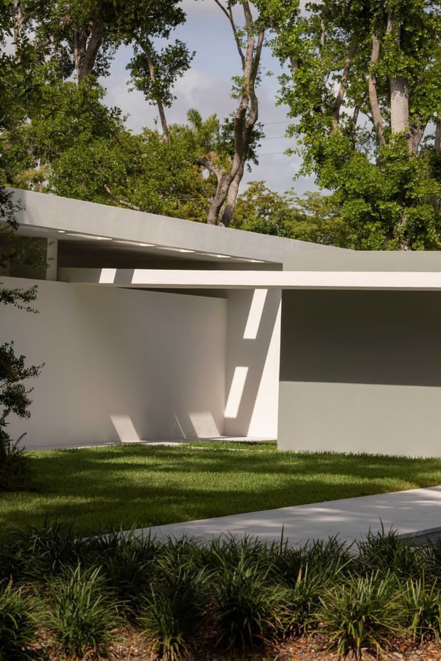 A minimalist contemporary white architectural exterior set within a mature landscaped garden. The low, flat-roofed structure features smooth white rendered walls, strong horizontal roof planes, deep overhangs, and angled structural supports that create sharply defined sunlit shadows. A lawn, paved walkway, and dense ornamental planting occupy the foreground, while mature trees rise behind the building.