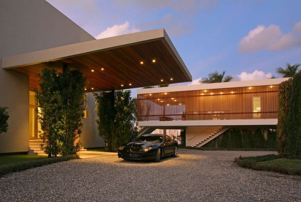 A modern residential exterior at dusk, featuring a large flat-roofed entrance canopy with warm timber soffit and recessed lighting, a raised upper-level wing with vertical timber screening and balcony, and an open external staircase. The home is approached via a broad gravel driveway with landscaped hedges and trees; a car is parked beneath the canopy. The composition emphasizes clean geometric forms, warm wood detailing, and indoor-outdoor architectural lighting.