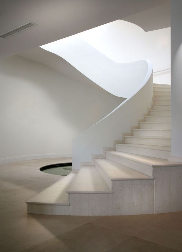 A sculptural interior stairway with a sweeping curved white balustrade and broad pale stone or tile treads. The staircase rises beside a softly lit architectural void or skylight, with minimal white walls, concealed lighting, and a small dark inset feature at the lower level. The design is highly refined and minimalist, emphasizing flowing geometry and clean surfaces.