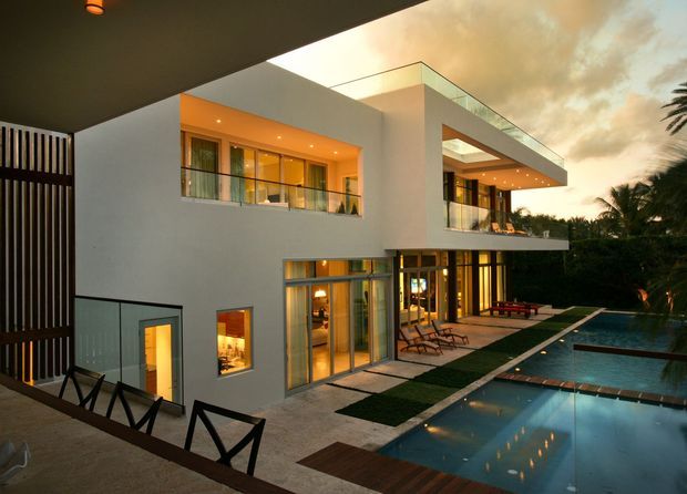 A modern multi-level residential home viewed from an elevated outdoor terrace, featuring crisp white cubic volumes, broad glazed openings, cantilevered balconies, a flat roof, and warm interior lighting at dusk. A long rectangular swimming pool, paved terrace, loungers, and landscaped tropical planting occupy the foreground and right side.