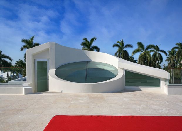 A low, sculptural white modern building facade under a vivid blue sky, featuring a large oval glazed opening, narrow horizontal glazing, and a flat, gently curved roofline. A broad pale stone or concrete terrace extends across the foreground, with tropical palm trees visible behind the building and a red foreground surface at the bottom edge.