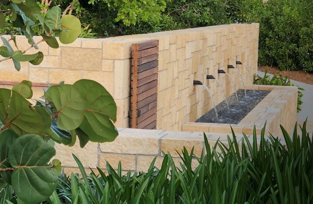 Exterior landscaped courtyard feature with a long shallow water fountain integrated into pale stacked stone walls. The wall includes a vertical dark timber gate or panel, while dense tropical planting frames the foreground and background.