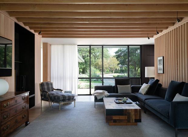 A sophisticated living room with a large dark sectional sofa, plaid lounge chair, low timber coffee table, and timber dresser. Full-height black-framed glazing opens views to a garden, while a warm exposed timber beam ceiling and vertical timber wall battens add architectural character. The room has a calm, refined, mid-century-influenced atmosphere.