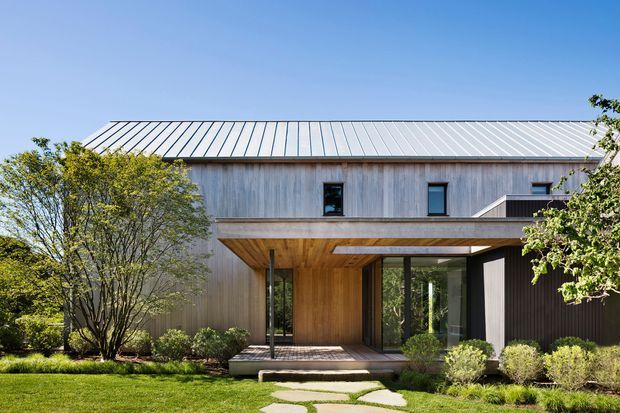 A contemporary two-storey residential house viewed from the landscaped front garden. The home features a broad low-pitched standing-seam metal roof, vertical weathered timber cladding, dark-framed windows, and a deep timber-lined entry canopy projecting over a recessed glazed entrance. Mature trees, clipped shrubs, lawn, and stepping-stone paving frame the facade.