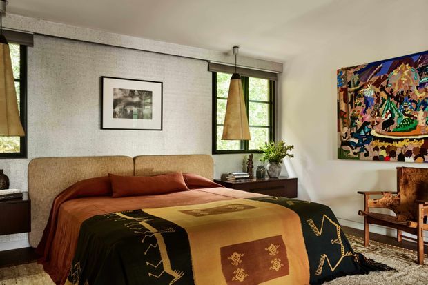 A warm, richly layered bedroom with a large upholstered bed positioned against a textured feature wall. The room has dark-framed windows with woven pendant shades, a low dark timber bedside cabinet, framed artwork, colourful abstract wall art, and a vintage-style timber lounge chair. Earthy rust, ochre, brown and black textiles create a cosy, eclectic atmosphere.