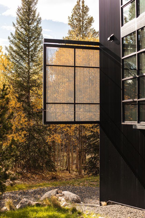 Exterior architectural photograph of a contemporary dark-clad building set within a wooded landscape. The facade features vertical ribbed cladding, large black-framed windows, and a projecting glazed box-like window or balcony enclosure. Autumn trees are reflected in the glazing and surround the structure, with gravel landscaping and rocks at ground level.