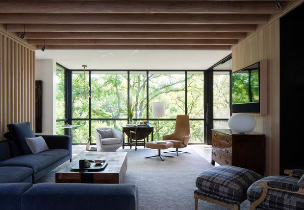 A warm, mid-century-inspired living room with a dark blue sectional sofa, lounge chairs, timber coffee table, low timber drawers, and a large rug. Full-height black-framed glazing opens the room visually to leafy trees, while exposed timber ceiling beams and vertical timber wall panelling add architectural warmth.