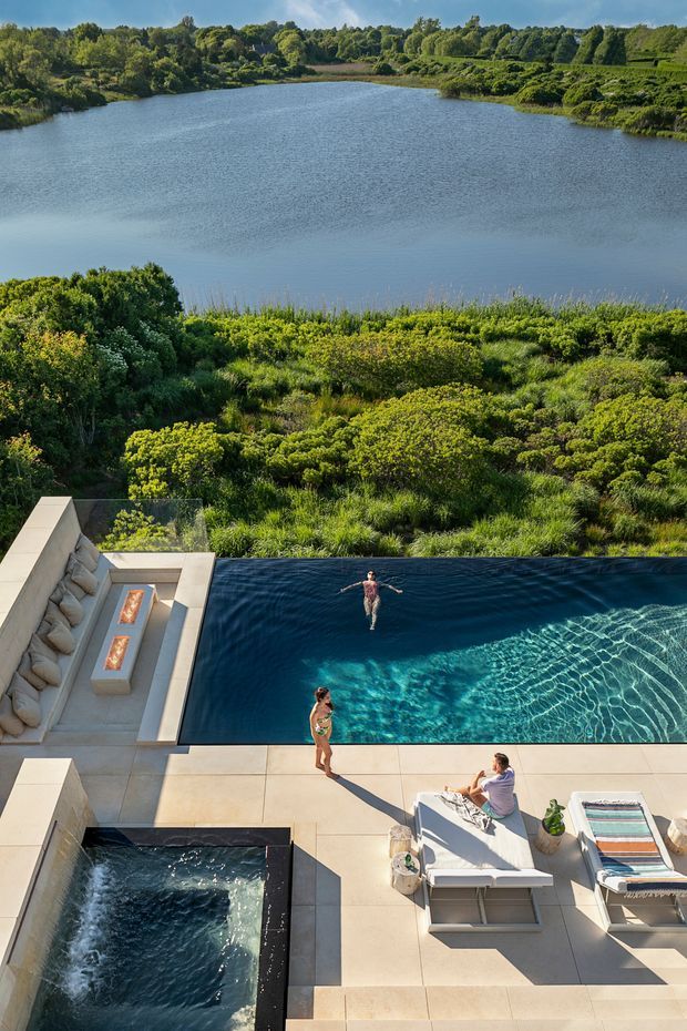 Elevated view of a luxurious outdoor pool terrace overlooking a calm lake and dense green landscape. The scene includes a long swimming pool, a separate square spa or plunge pool, pale stone paving, built-in lounge seating with fire features, sun loungers, and people using the water and terrace. Bright natural light and expansive water views create a resort-like setting.