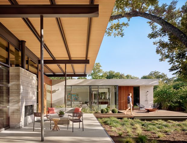 A modern single-storey residential courtyard or patio viewed from beneath a broad covered overhang. The composition features extensive glazing, slender dark structural posts, warm timber soffits and cladding, pale masonry walls, concrete paving, timber decking, and landscaped planting surrounding an outdoor seating area. A low flat-roofed pavilion is visible beyond the courtyard.