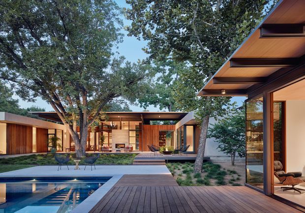 A contemporary single-storey residence arranged around a landscaped courtyard and swimming pool. The image shows extensive timber decking, warm timber-clad walls and soffits, broad glazing, low horizontal roof forms, and an open indoor-outdoor living arrangement surrounded by mature trees.