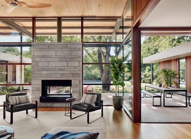 A modernist living room with a central grey masonry fireplace, low black-framed lounge chairs, and extensive floor-to-ceiling glazing overlooking mature trees. Warm timber flooring, a timber-lined ceiling with ceiling fan, exposed structural posts, and dark timber window framing create a strong indoor-outdoor connection. Through the open glazed wall is a covered outdoor dining area with a long table and chairs.