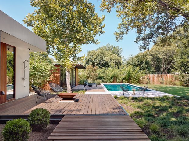 A landscaped backyard with a modern low-profile home opening onto a broad timber deck and garden terrace. The deck includes outdoor seating and a central fire feature, with a rectangular swimming pool beyond, surrounded by pale paving, mature trees, lawn, and naturalistic planting. The architecture features clean horizontal lines, rendered walls, extensive glazing, and warm timber screening.