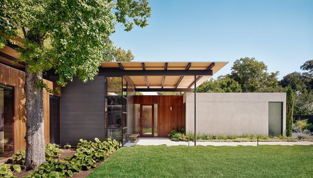 A contemporary single-storey residential facade viewed from the landscaped front lawn. The design combines dark timber cladding, a broad flat projecting canopy with exposed slender framing, extensive glazing, a warm timber entry, and a simple pale rendered or concrete side volume. Mature trees, lawn, and planting surround the building.