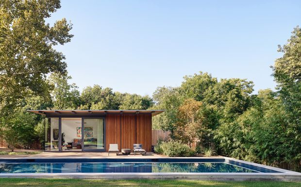 A compact modern poolside pavilion or backyard studio set among mature trees and dense landscaping. The building has a low, flat projecting roof, extensive glazing on the front, warm vertical timber cladding, and dark slender framing. A long rectangular swimming pool occupies the foreground, reflecting the structure and surrounding greenery.