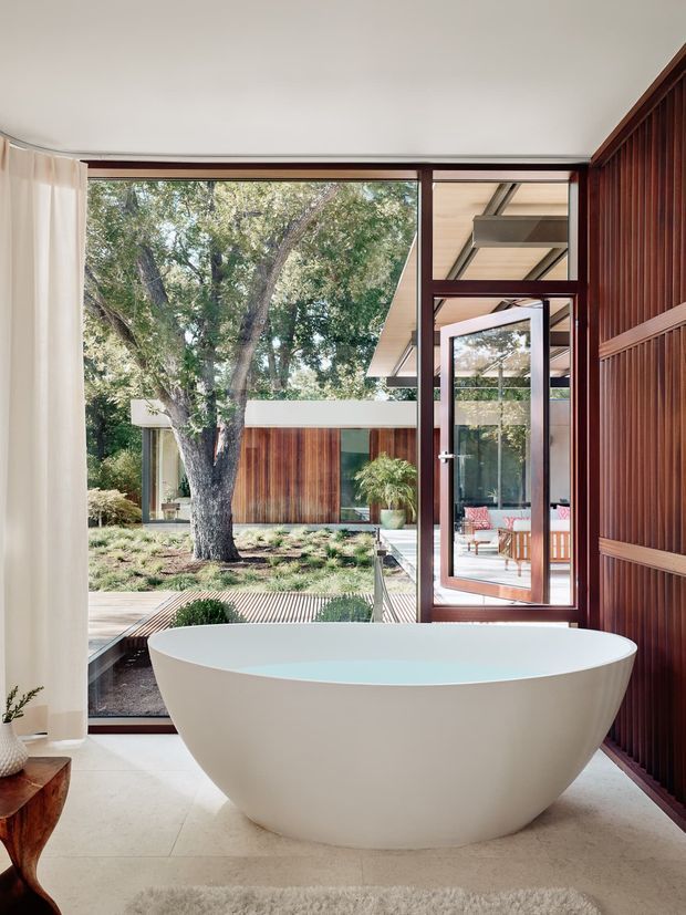 A serene, contemporary bathroom centered on a freestanding oval white bathtub. Full-height glazing and a glass door open toward a landscaped courtyard, while dark timber vertical panelling frames the right wall. Pale stone or tile flooring, sheer curtains, warm timber detailing, and abundant views of mature trees create a calm indoor-outdoor connection.