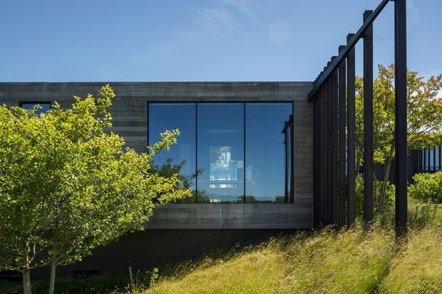 Exterior view of a contemporary low-profile building set into a gently sloping landscaped site. The facade combines dark horizontal cladding or concrete-like panels with a large floor-to-ceiling glazed window, framed in dark metal. A tall black vertical screen or fence runs along the right side, with mature shrubs and grasses integrated into the foreground landscaping.